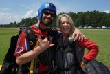 Two people in skydiving gear stand on a grassy field. They are smiling and posing with arms around each other, with one person making a hand gesture. Both are wearing helmets and parachute harnesses. A line of trees and a blue sky are in the background.