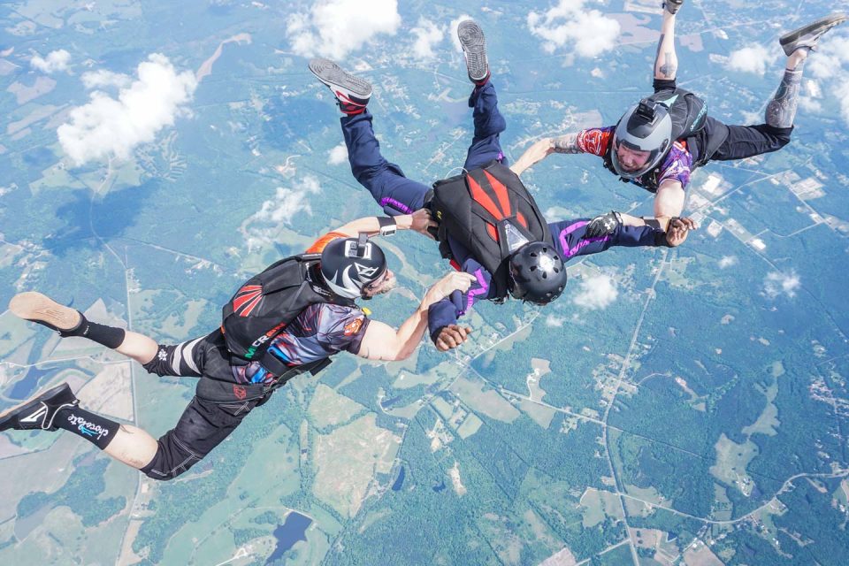 Three skydivers in formation hold hands mid-air against a backdrop of clouds and landscape far below. They're wearing colorful gear with helmets and backpacks.