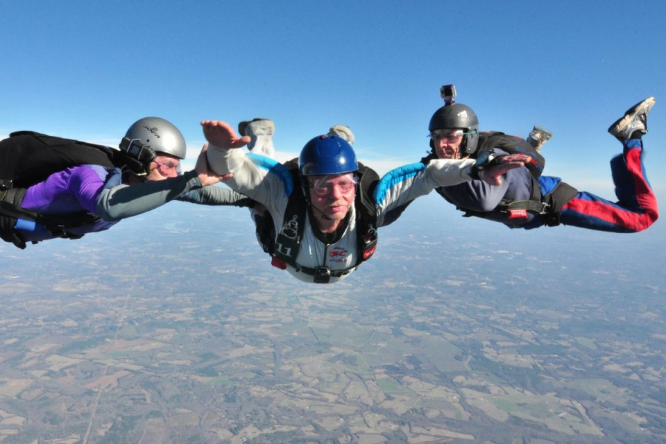 Three skydivers in a freefall formation above a vast landscape of fields and forests. They are wearing helmets and jumpsuits and appear to be enjoying the clear, sunny day.