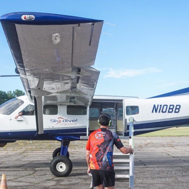 A man wearing a colorful shirt climbs a small set of stairs leading to a blue and white propeller plane with "Skydive Carolina" branding. The plane is parked on a tarmac with trees and a clear blue sky in the background.