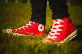 A person wearing red high-top Converse sneakers stands on a grassy field. The shoes have white laces and a circular logo on the side. The person is wearing black pants.