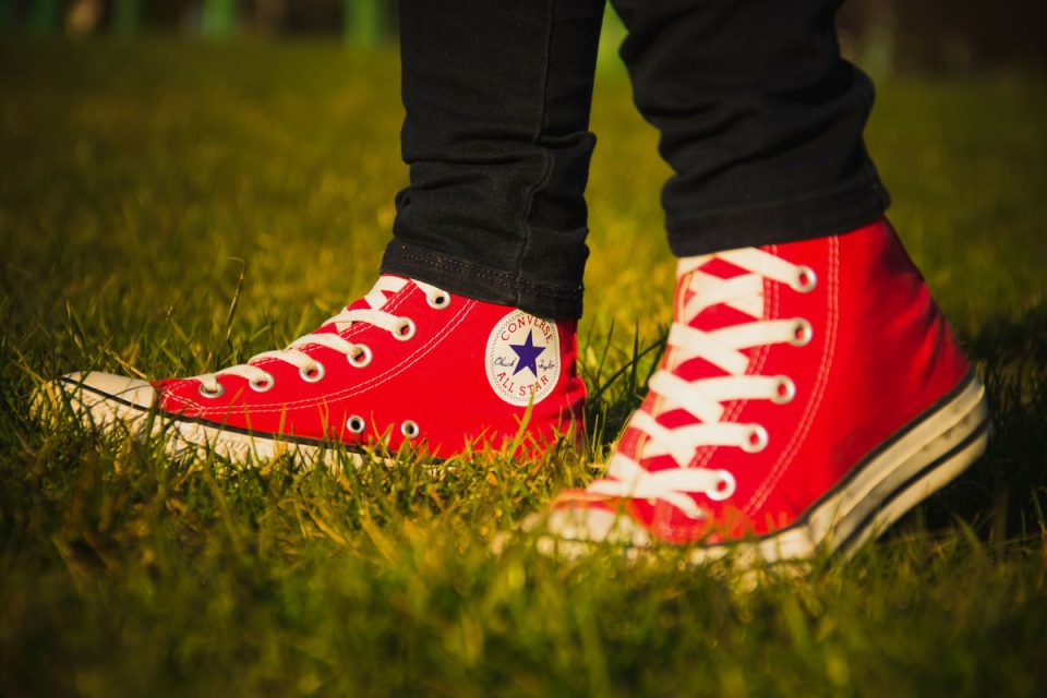 A person wearing red high-top Converse sneakers stands on a grassy field. The shoes have white laces and a circular logo on the side. The person is wearing black pants.