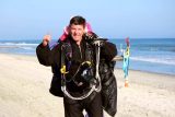 A person wearing a parachute harness and suit, smiling and giving a thumbs-up on a sandy beach. The ocean is visible in the background on a clear, sunny day. Multicolored flags are also present along the shore.