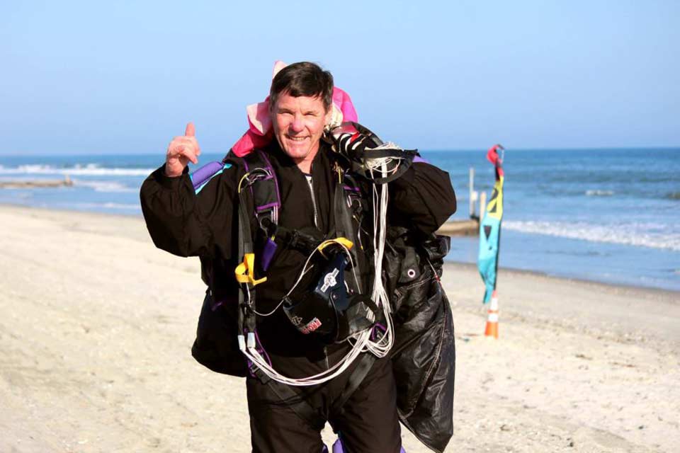 A person wearing a parachute harness and suit, smiling and giving a thumbs-up on a sandy beach. The ocean is visible in the background on a clear, sunny day. Multicolored flags are also present along the shore.