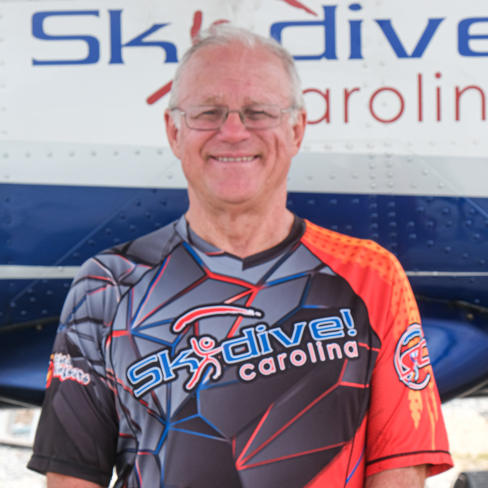 An older man wearing glasses and a Skydive Carolina shirt stands smiling in front of an airplane. The shirt is predominantly gray with red and orange accents.