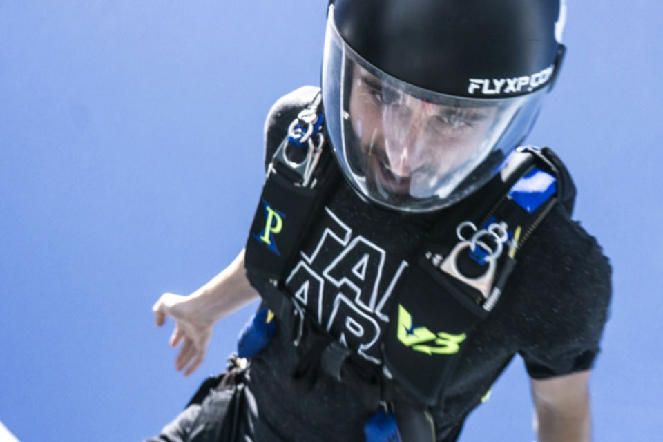 A person skydiving in a clear blue sky, wearing a helmet and a harness. The individual has a focused expression and a shirt with colorful text.