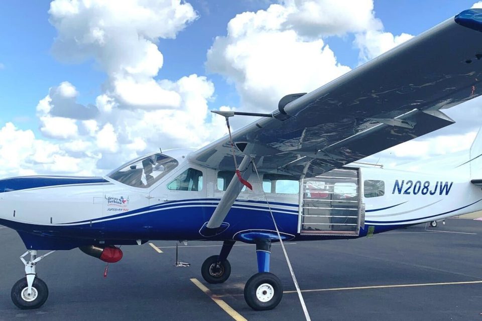 A small white and blue single-propeller plane is parked on an airport tarmac under a partly cloudy sky. The tail number N208JW is visible. The passenger door is open, showing a glimpse of the interior.