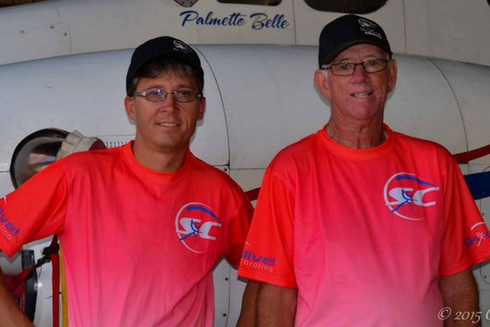 Two men wearing matching bright pink shirts and black caps stand in front of an airplane. Both are smiling and appear to be in a hangar. The airplane has "Palmetto Belle" written on it.