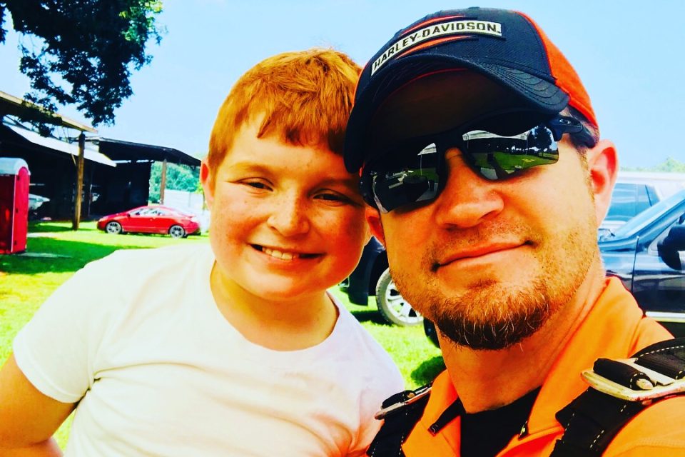 A man wearing sunglasses and a cap smiles while taking a selfie with a young boy in a white shirt. They are outdoors on a sunny day, with cars and trees visible in the background.