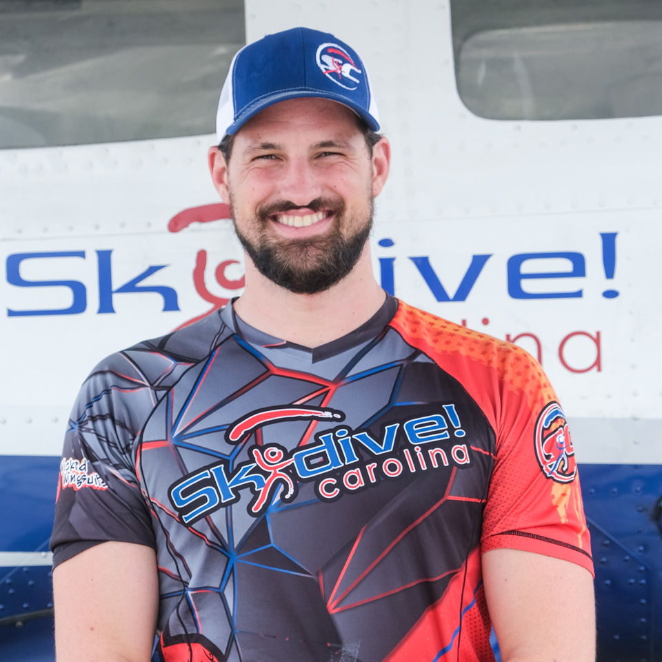 A person smiling and standing in front of a Skydive Carolina plane. They are wearing a colorful Skydive Carolina shirt and a cap.