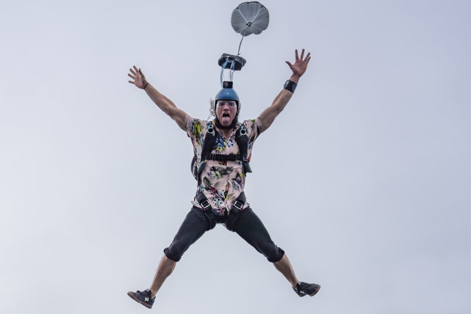 A person skydiving with a parachute deployed, wearing a floral shirt, black pants, and a blue helmet, is captured mid-air against a clear sky, with arms and legs extended outward.