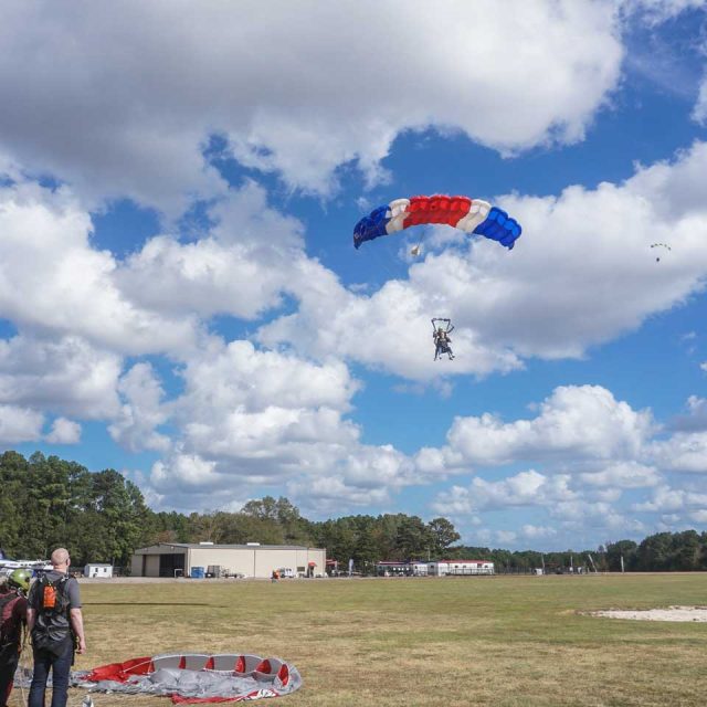 A paraglider with a blue, white, and red parachute descends towards an open grassy field. Two people stand nearby, looking up at the sky. A few structures and trees are visible in the background under a sky dotted with clouds.