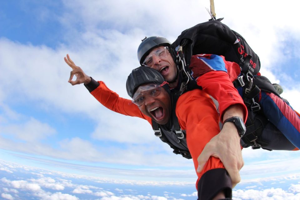 Two people are skydiving together, wearing helmets and safety gear. The person in front has their mouth open in excitement and is giving an "OK" gesture. They are above the clouds with a blue sky backdrop.