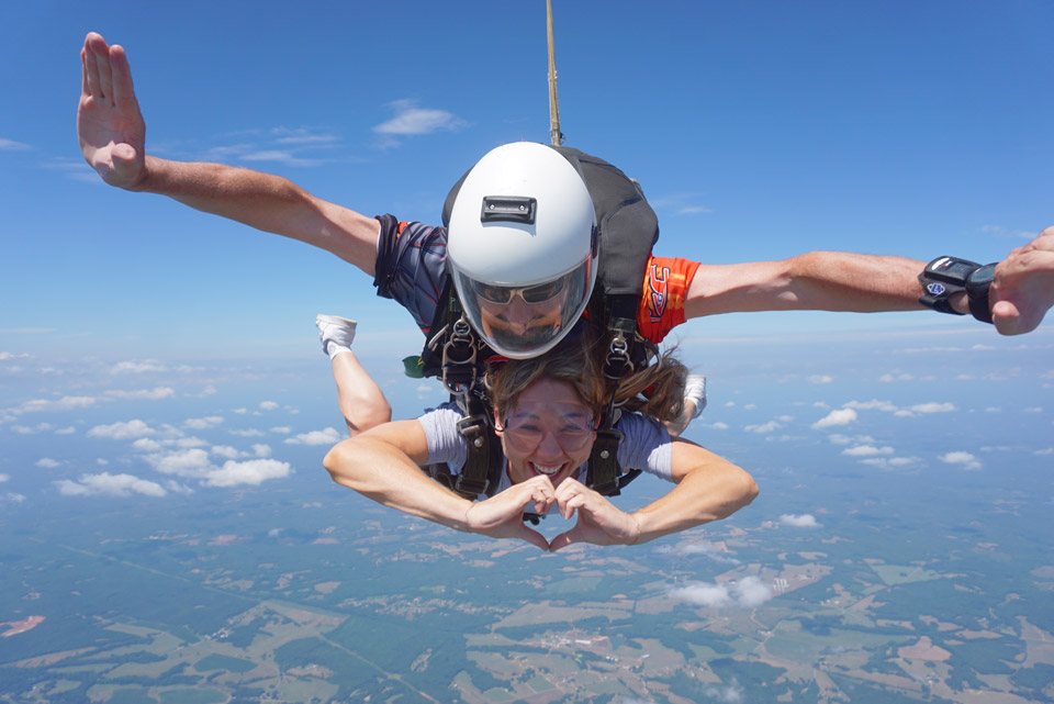 Two people tandem skydiving against a backdrop of a blue sky and scattered clouds. The person in front is making a heart shape with their hands, while the instructor behind manages the parachute harness. Below, a patchwork landscape is visible.