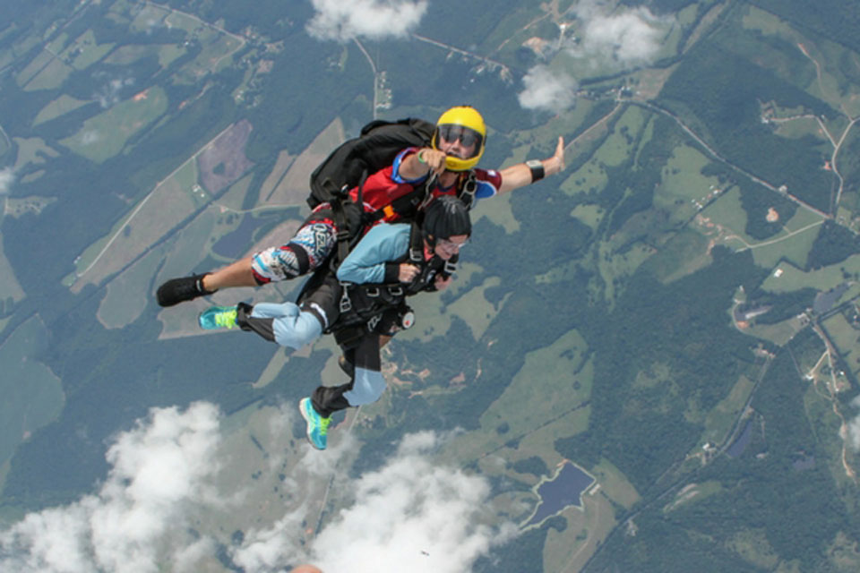 A tandem skydiver and an instructor in mid-air above a landscape of fields and forests. The instructor wears a yellow helmet and gestures with excitement. The skydiver is dressed in a light blue outfit. White clouds are scattered around them.