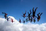 A group of skydivers performs an aerial formation against a backdrop of clear blue sky and fluffy white clouds. They are spread out in various poses, wearing colorful jumpsuits and helmets.