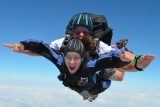 Two people tandem skydiving in mid-air, both wearing helmets and goggles. The person in front is smiling excitedly, while the instructor is behind, guiding the descent. The bright blue sky and clouds form the background.