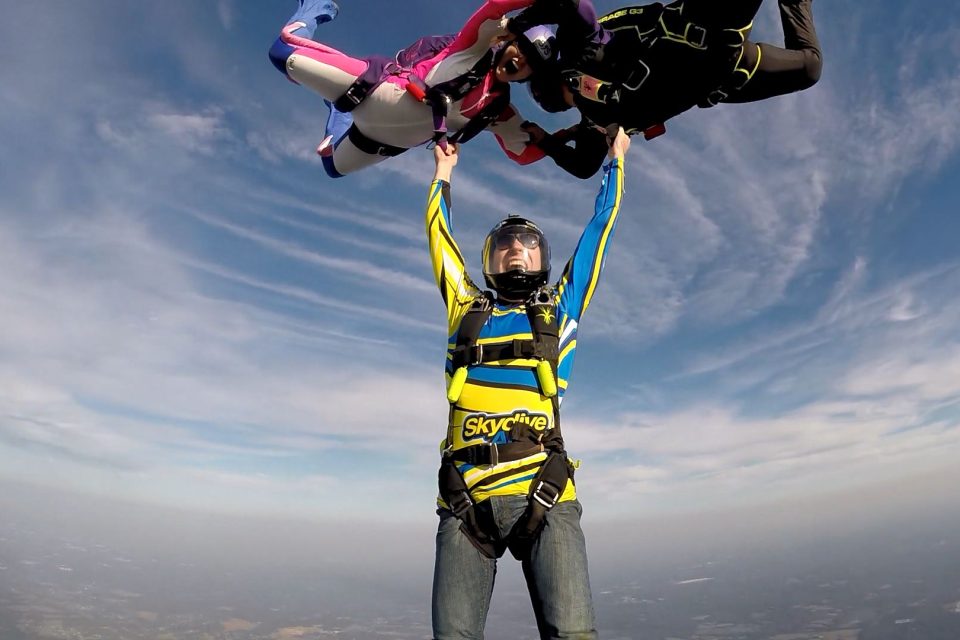 Three skydivers are mid-air against a backdrop of blue sky and wispy clouds. Two are wearing vibrant suits and helmets, holding onto a third person who is upright below them. The scene captures them during a dynamic skydive maneuver.