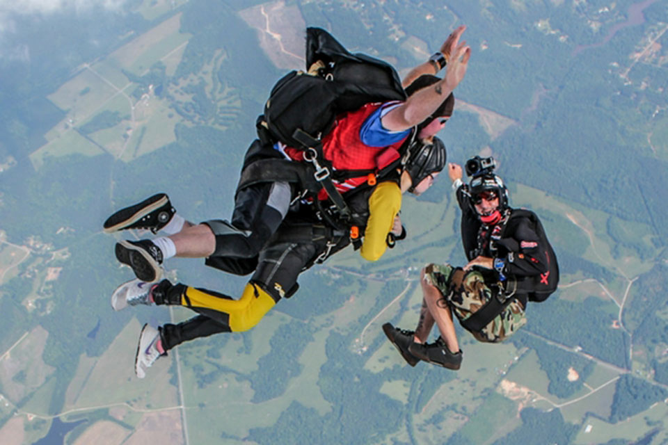 Two people in a tandem skydive, one wearing a red shirt and yellow pants, soar through the air with a cameraman skydiver capturing the moment. They are above a landscape of fields and forests, and are in free fall mid-air.