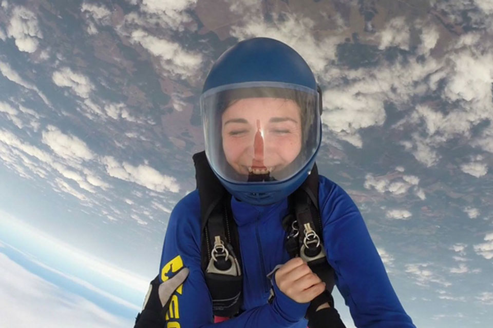 A person with a blue helmet and suit is skydiving, smiling broadly as they descend through a sky filled with fluffy clouds. The ground is visible far below.