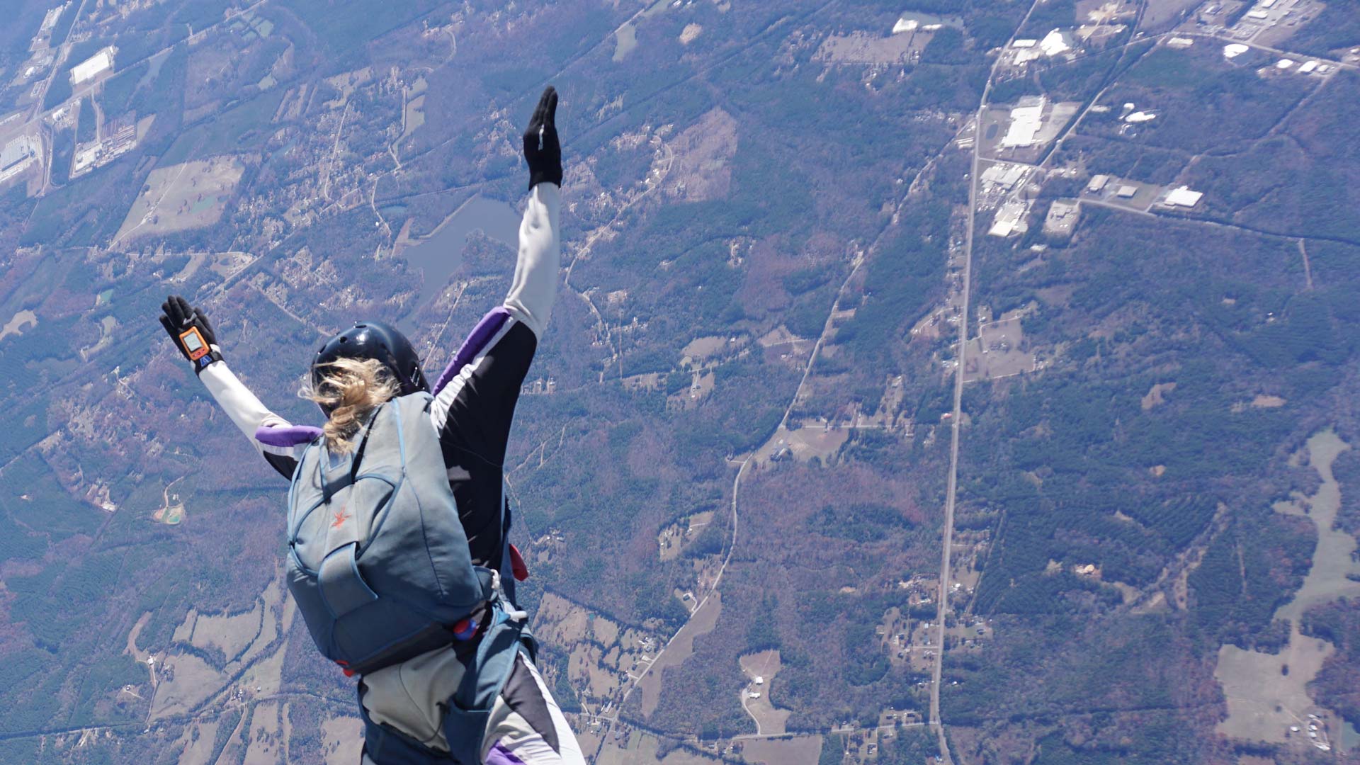 A skydiver wearing a gray and black jumpsuit and helmet, posing mid-air with one arm raised against a backdrop of a rural landscape, including fields, roads, and scattered buildings.