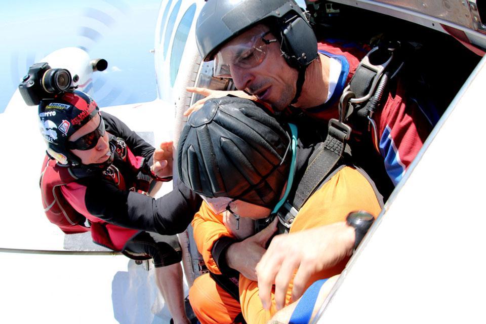 Three skydivers prepare to jump from a plane. One wears an orange jumpsuit and black helmet, another has a red suit and black headwear. The third, assisting from inside the plane, wears a blue and red outfit. All are focused and equipped with helmets.