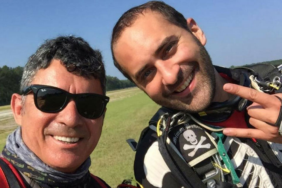 Two men in skydiving gear smiling at the camera. One wears sunglasses and the other holds up a peace sign. They stand on a grassy field under a clear blue sky.