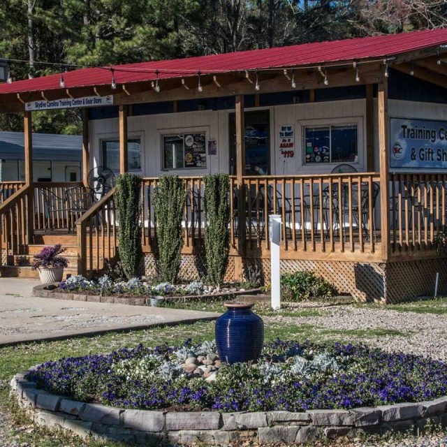 A wooden building with a red roof features a porch and signs that read "Training Center" and "Gift Shop." In front, there is a circular flower bed with purple flowers and a decorative blue vase. Trees are visible in the background.