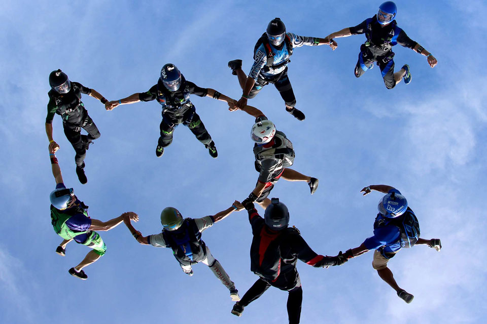 A group of eight skydivers in mid-air, forming a circle as they hold hands against a clear blue sky. They wear colorful jumpsuits and helmets, creating a dynamic and synchronized formation.