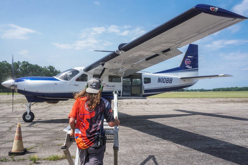 A person with long hair wearing a bright shirt and cap walks toward a small blue and white airplane on an airport runway. The sky is clear, and there are trees in the background. The aircraft has "N1088" marked on its side.