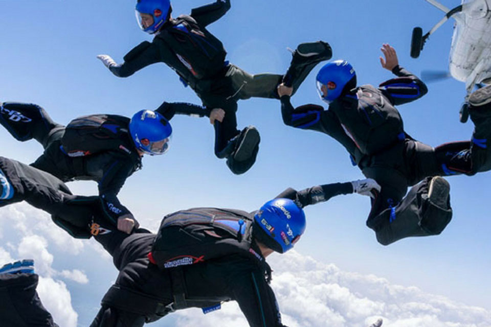 Five skydivers wearing blue helmets and black jumpsuits perform a synchronized formation against a backdrop of clear blue sky and clouds. An airplane can be seen in the top right corner.