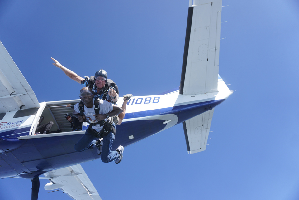 Two people are tandem skydiving, jumping out of a small aircraft. The sky is clear and blue, and they are harnessed together, with the instructor positioned above the other person, both appearing excited.