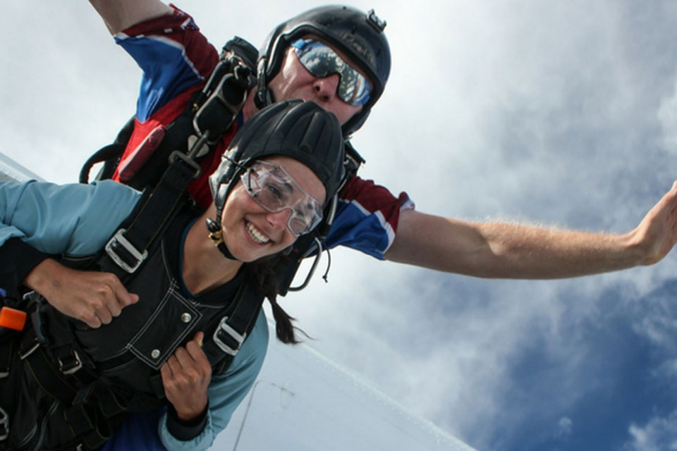 A person tandem skydiving with an instructor. They are both wearing helmets and goggles, with parachute gear secured. The sky is blue with some clouds in the background. The person in front is smiling broadly, holding out their arms.