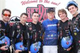 A group of six skydivers dressed in black jumpsuits, five holding blue helmets, stand together smiling in front of a Skydive Arizona sign. They appear to be posing for a group photo.