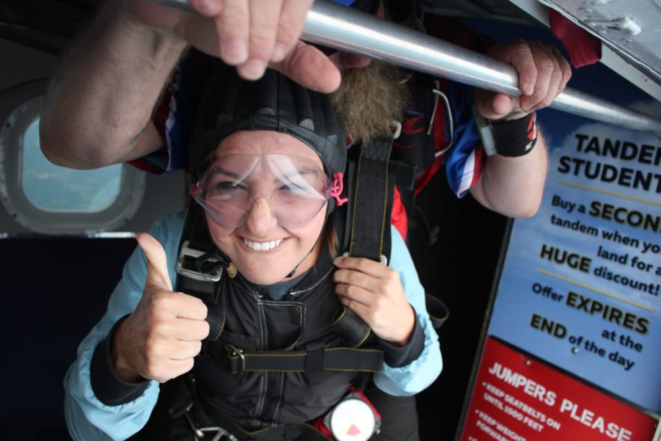 A person wearing skydiving gear gives a thumbs-up while standing at the open door of an aircraft, ready for a tandem jump. An instructor is visible in the background behind them. A sign with text is partially visible to the right.