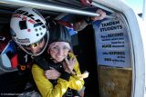 A tandem skydiving instructor and a smiling woman prepare to jump from an airplane. Both wear helmets and parachute gear. A sign inside the plane offers discounts for additional jumps.