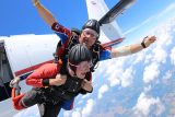 Two people tandem skydiving from a plane, wearing helmets and parachute gear. The instructor is guiding the other person, and both are in free fall above a landscape with clouds below them. The sky is bright and clear.