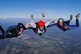 Three skydivers in free fall, wearing jumpsuits and helmets, link arms high above a patchwork of fields and forests visible below against a clear blue sky.