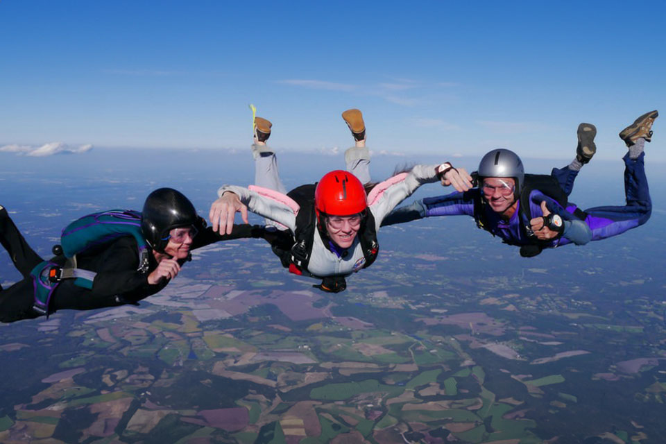 Three skydivers in free fall, wearing jumpsuits and helmets, link arms high above a patchwork of fields and forests visible below against a clear blue sky.