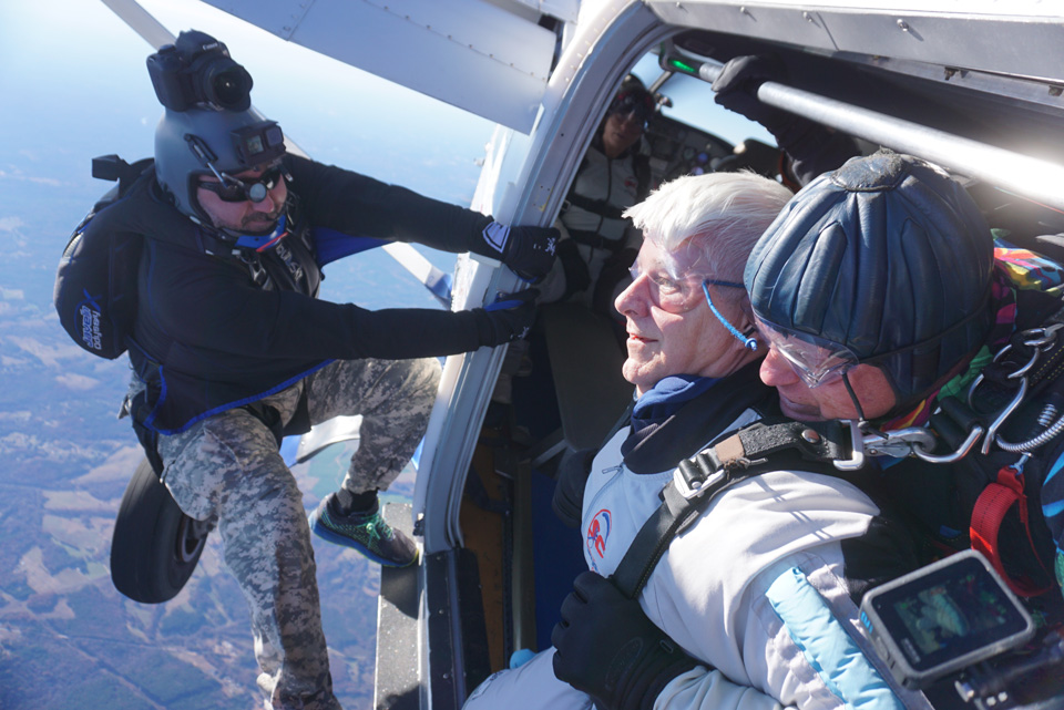 Two skydivers, one older and one younger, prepare to jump out of an airplane. The younger skydiver wears a helmet camera and assists the older man, who wears goggles. A third person is visible inside the aircraft. The landscape below is clear and distant.