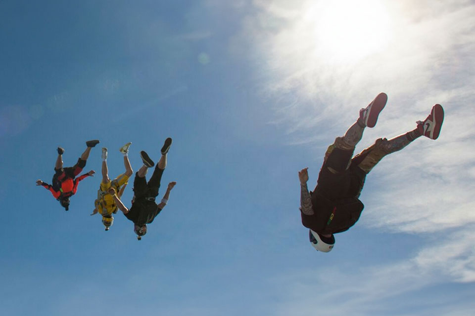 Four skydivers in colorful outfits perform a formation jump against a clear blue sky. The sun is shining brightly, creating a glowing effect. The skydivers are upside down, showcasing an impressive aerial maneuver.