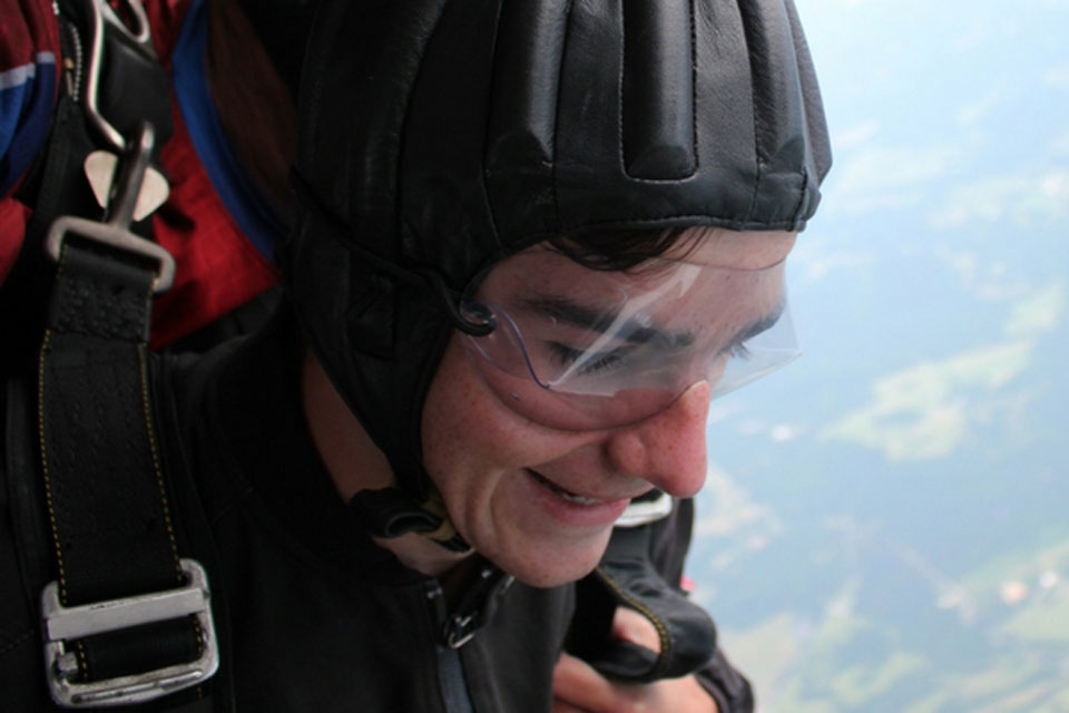 A person wearing a black helmet and clear goggles is skydiving, with a parachute harness visible. They are smiling as they descend, with a landscape of fields in the blurred background.