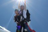 Two people are tandem skydiving, smiling and holding onto each other as they jump from a plane. The sky is clear and blue, with the sun shining brightly above them. They are wearing helmets and parachute gear.