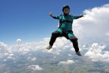 A person in a teal jumpsuit and helmet is skydiving, arms outstretched against a backdrop of blue sky and white clouds. The landscape below features fields and patches of greenery.