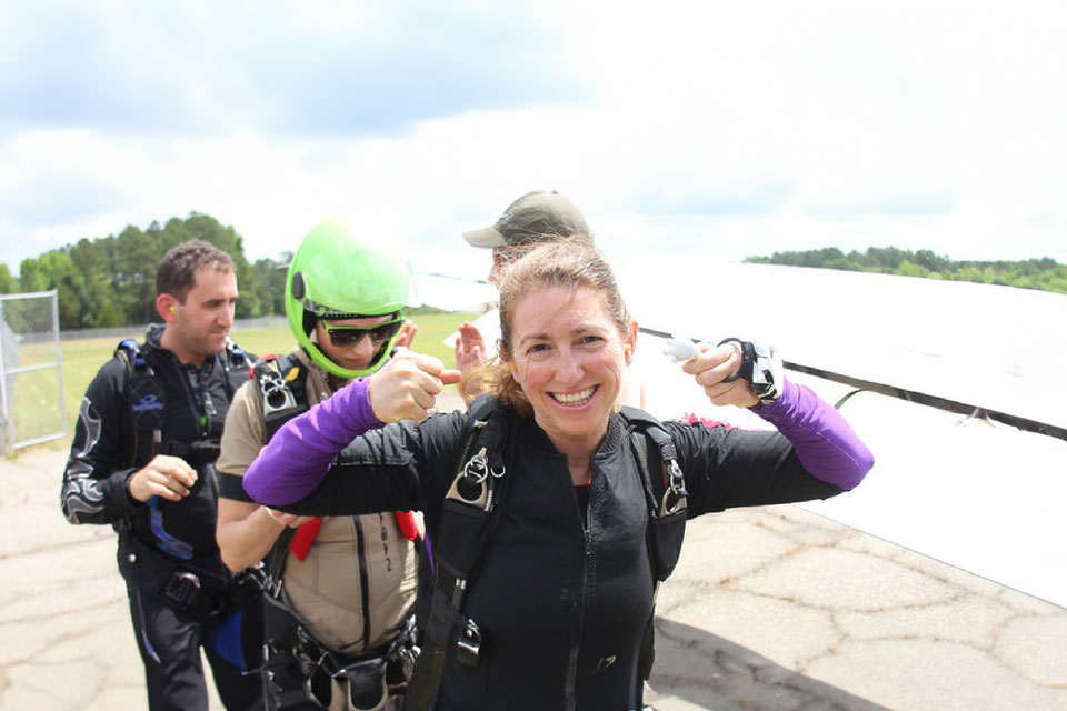 A woman smiles and gives thumbs up while wearing skydiving gear. Two other individuals in skydiving suits and helmets stand nearby. The background shows an airfield with trees and a cloudy sky.