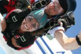 A person tandem skydives with an instructor, both wearing helmets and goggles. They are exiting a plane mid-air. The person in front is smiling widely, showing excitement, with a blue sky in the background.