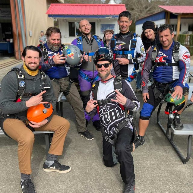 A group of eight skydivers in jumpsuits and harnesses pose together outdoors, some holding helmets. They're sitting and kneeling on benches in a recreational area with buildings in the background. Everyone is smiling and looking at the camera.