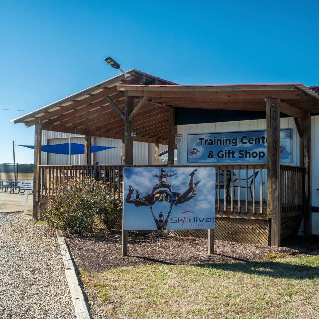 A wooden building with a sign reading "Training Center & Gift Shop" is seen. A large poster of skydivers is displayed in front, labeled "Skydive Carolina." The area is surrounded by gravel and plants under a clear blue sky.