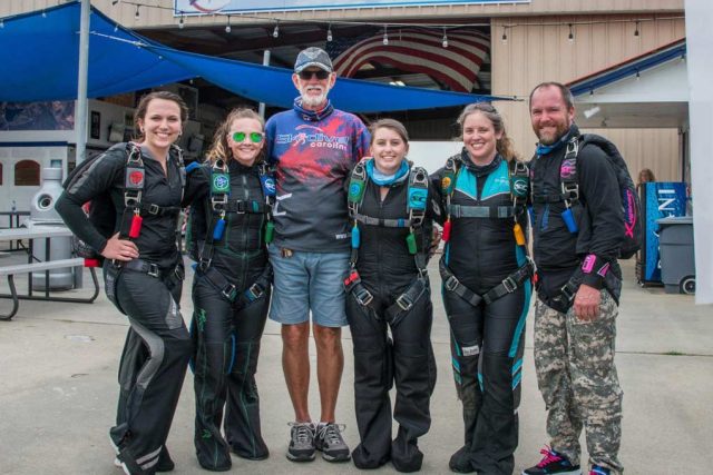 A group of five people in black skydiving suits and one person in casual clothes stand smiling together outside a building. The skydivers have colorful gear, and they all appear to be enjoying a sunny day.