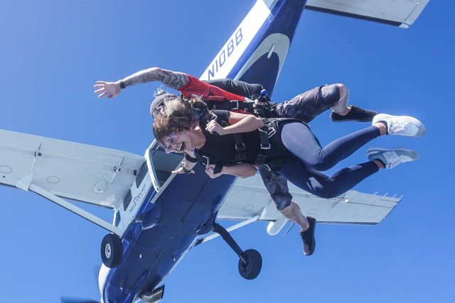 Two people skydive in tandem from a blue and white airplane against a clear blue sky. They appear excited and are in free fall with parachute harnesses. The aircraft is visible above them, and one engine propeller can be seen.